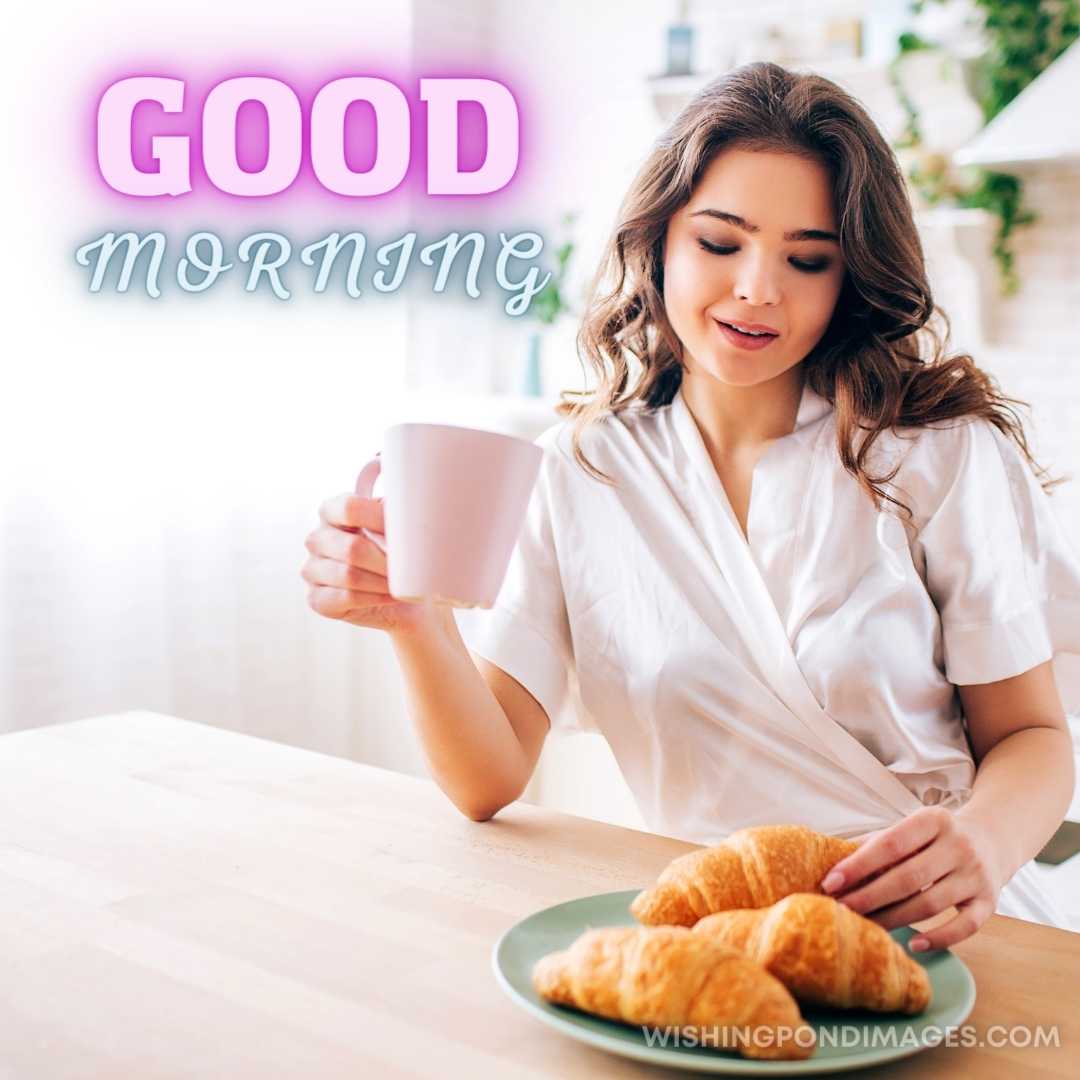 Young Woman with dark hair sitting in kitchen and drinking coffee in Morning taking one croissant an